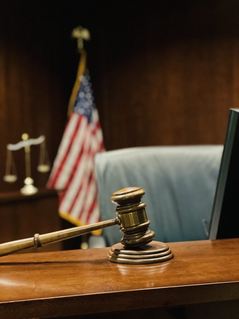 Selective focus on gavel, judge’s bench, scales of justice and flag in background.